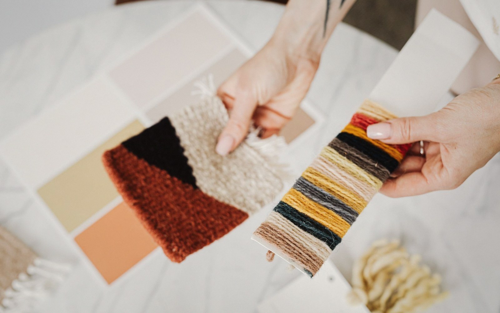 Hands compare woven fabric and yarn color swatches over paint chips on a table