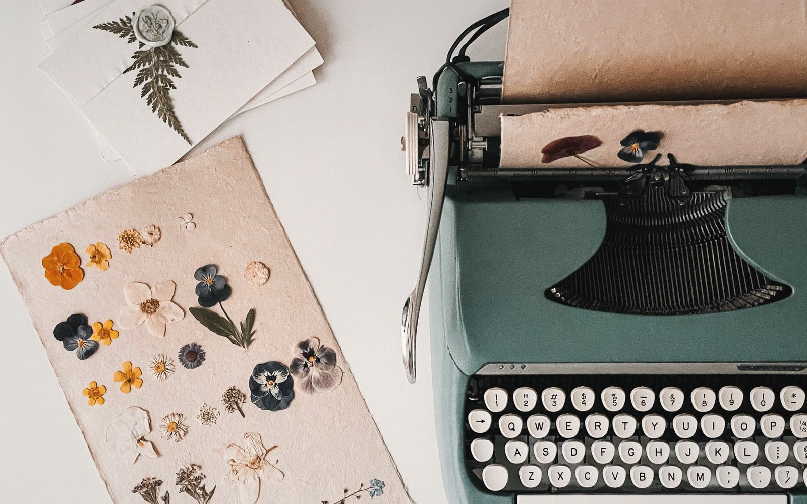 Vintage teal typewriter beside pressed flowers and fern arranged on paper