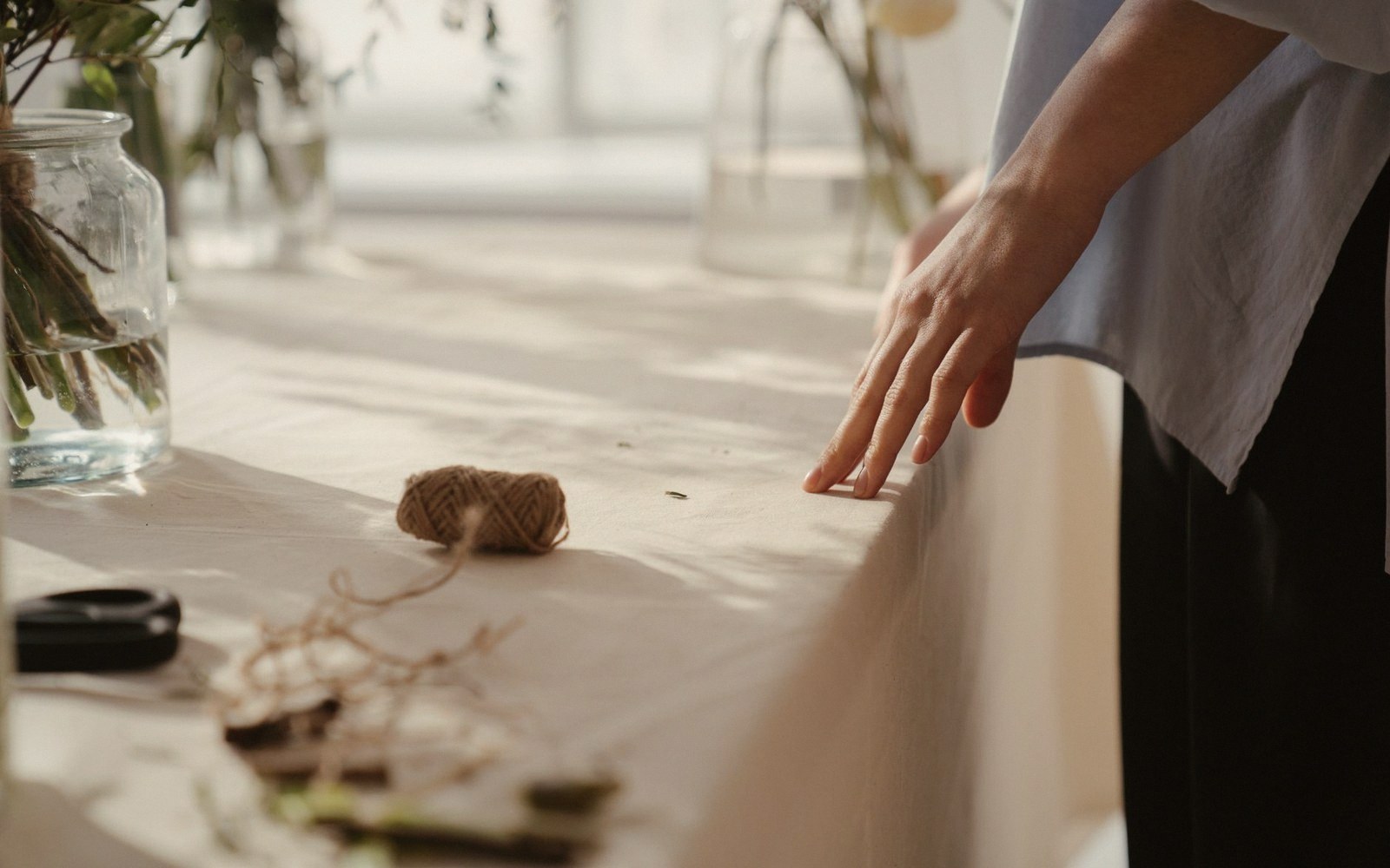A hand rests on a linen-covered table beside twine and glass jars of foliage